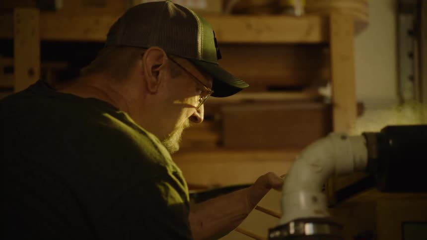 A senior woodworker with glasses and a cap meticulously sands wood in his dimly lit workshop
