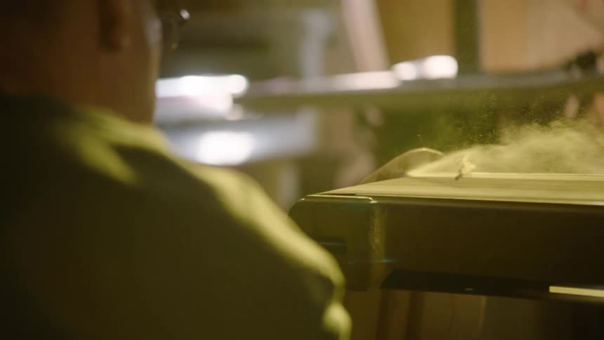 Cinematic slow-motion shot from over the shoulder captures a woodworker cutting a plank on a table saw as fine sawdust fills the air
