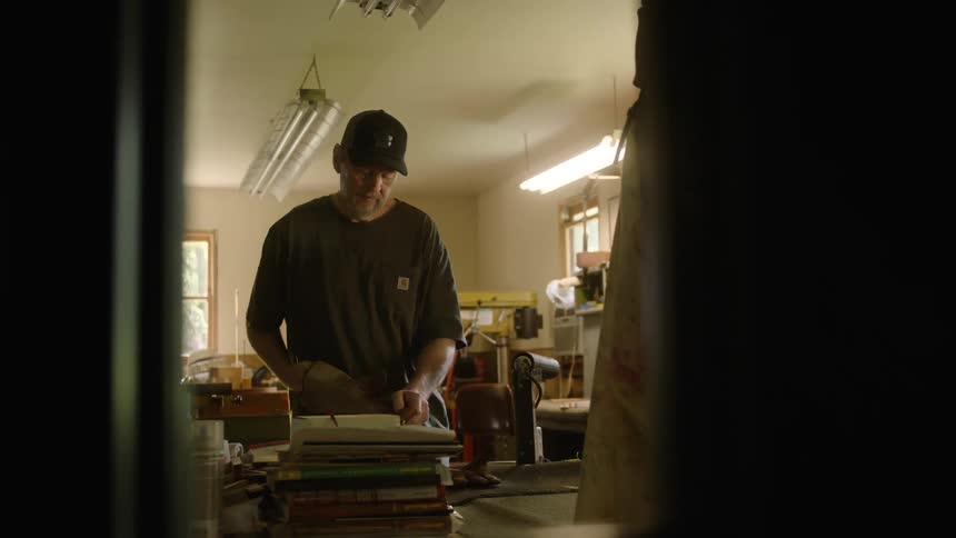 Viewed from a distance, a craftsman in a cap and work gloves carefully inspects and polishes a piece of leather in his workshop