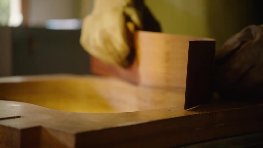 A craftsman wearing protective gloves carefully shapes a steam-bent piece of wood for a guitar body in a workshop