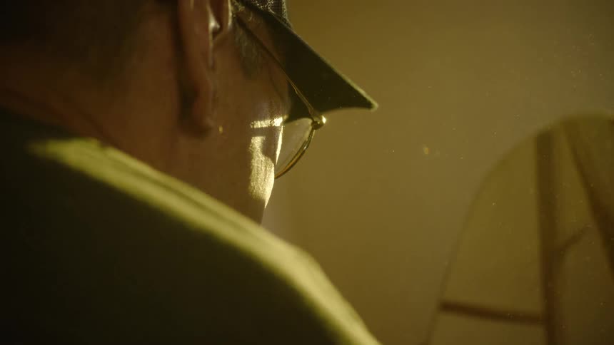A woodworker sands a piece of wood on a belt sander, with sawdust particles illuminated by warm light in slow motion