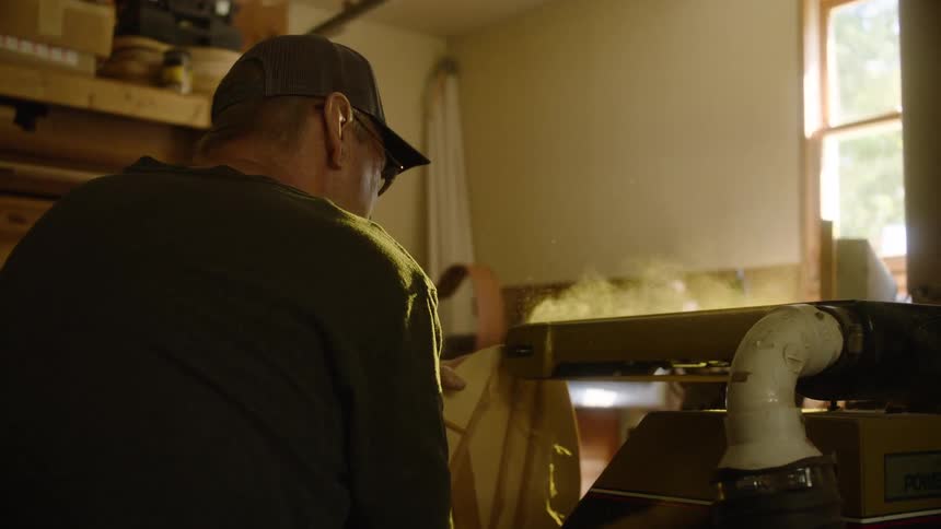 A woodworker skillfully sands a piece of wood on a belt sander in his workshop, with sawdust particles gracefully floating through the air in slow motion
