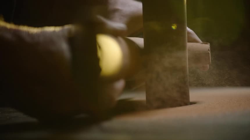 Close up of a craftsman's hands shaping wood on a vertical belt sander in slow motion, with sawdust particles glittering in the light