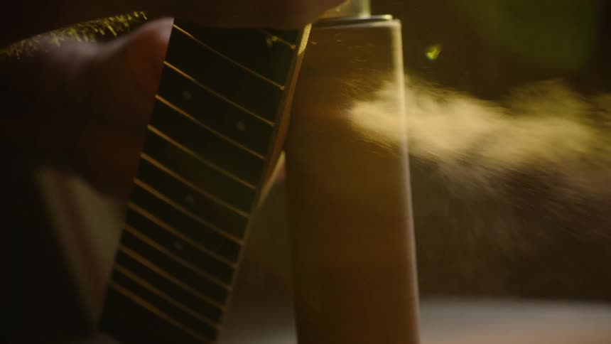 A luthier carefully applies a finishing spray to a guitar neck in slow motion, the fine mist illuminated by warm light