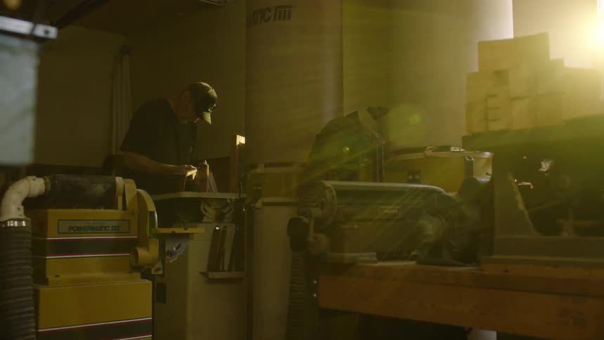 A focused craftsman sands a piece of wood on a belt sander in his workshop, backlit by a bright lens flare