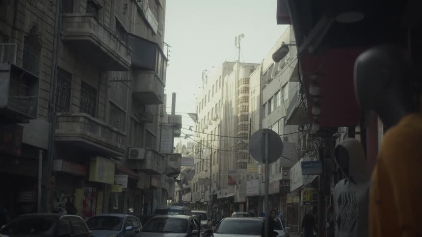 A static shot looking down a busy, narrow city street lined with shops and apartments on a bright day