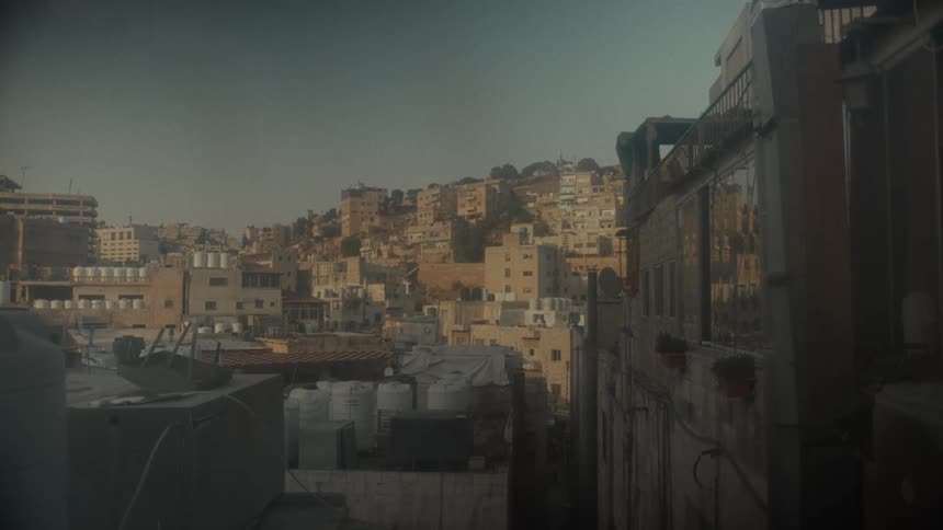 A sun-drenched, static view of a dense Middle Eastern city with buildings covering a hillside under a hazy sky