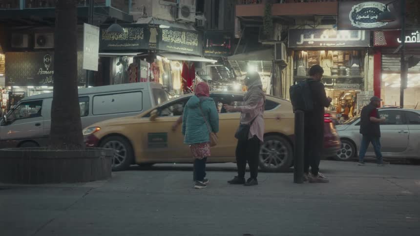 People walk and stand on a bustling city sidewalk at dusk as cars pass by brightly lit storefronts