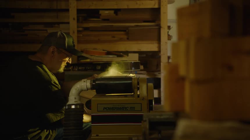 Focus pulls from a stack of wood to reveal a skilled luthier carefully sanding an acoustic guitar body in his workshop