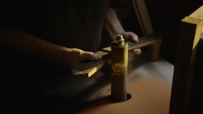 A craftsman in a dark workshop uses a power spindle sander to meticulously shape the neck of a guitar in dramatic slow motion