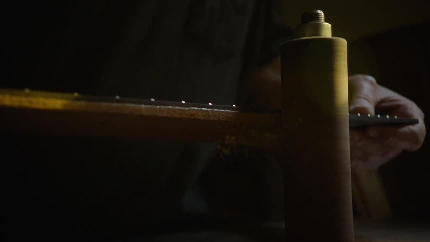 A craftsman sands the heel of a guitar neck with a spindle sander, sending sawdust flying in dramatic slow motion