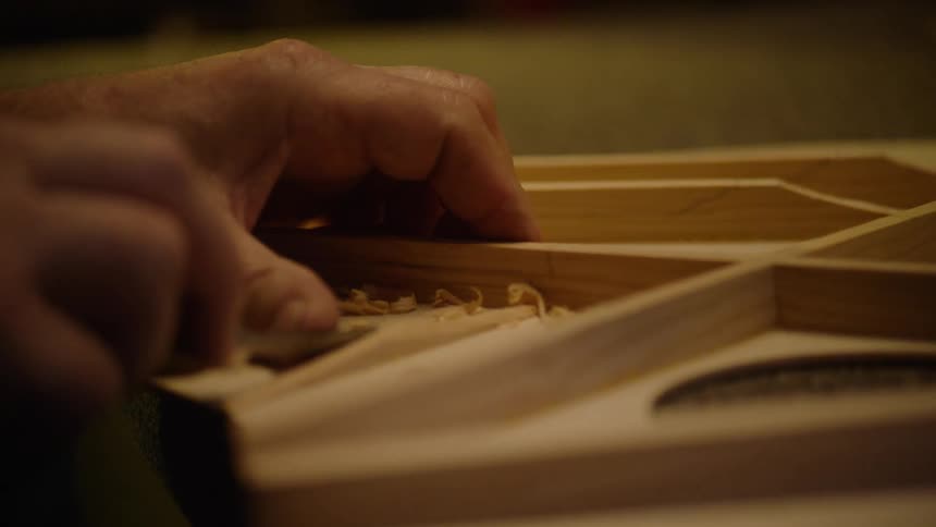 A skilled luthier's hands meticulously carve and shape the delicate wooden bracing for a custom acoustic guitar in slow motion