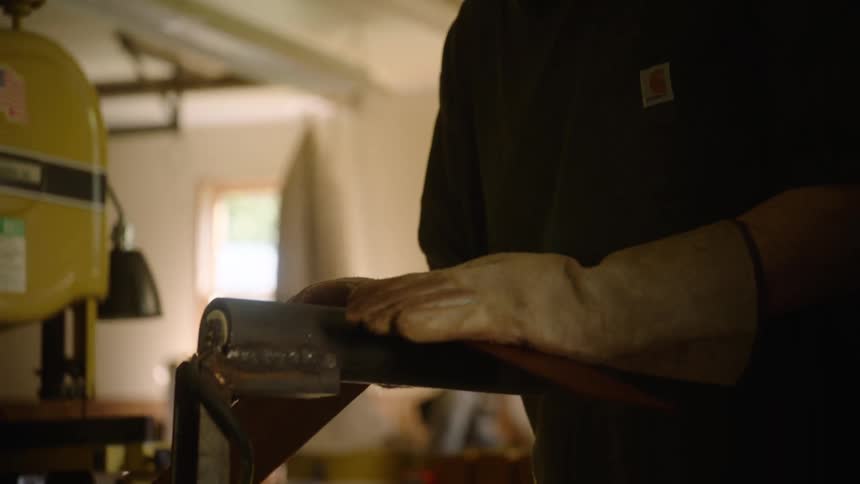 A focused craftsman carefully sands a metal knife blade on a belt sander in a dimly lit workshop