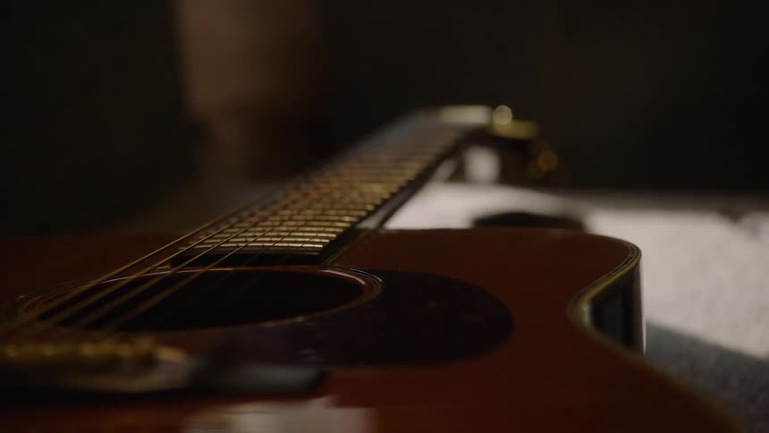 A dramatic close-up rack focus along the neck of a beautiful acoustic guitar in a dimly lit room