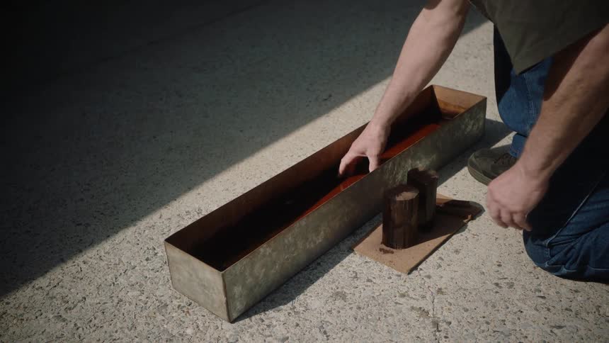 A carpenter kneels down to dip wooden blocks and a long board into a trough of dark stain in slow motion