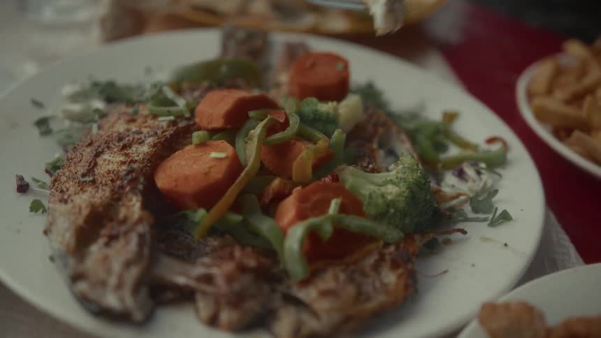 A fork lifts a tender piece of fish from a plate of grilled fish and vegetables in slow motion