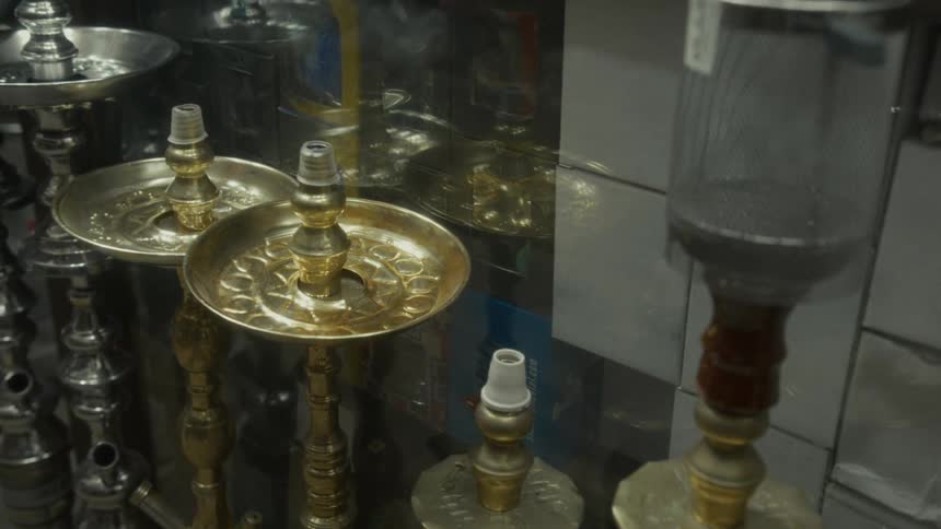 A static shot shows a collection of ornate gold and silver hookahs displayed in a shop window