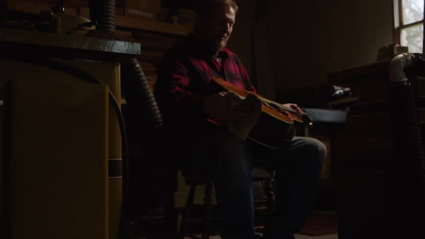 A craftsman sits in his dimly lit workshop and carefully cleans his prized acoustic guitar
