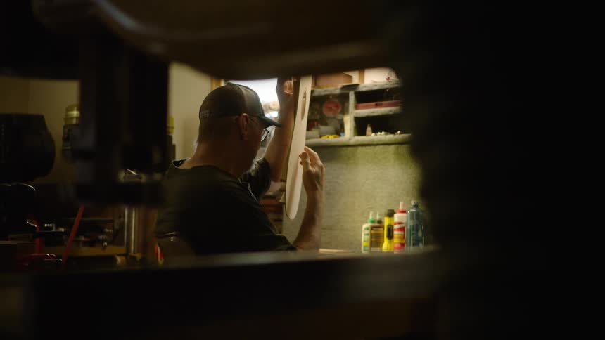 A luthier carefully inspects the soundboard of a guitar he is building in his dimly lit workshop
