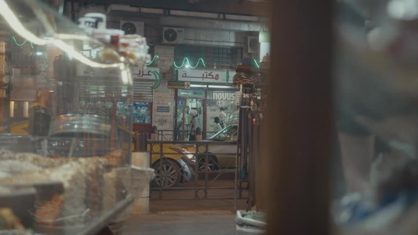 Looking through a shop window at a bustling Middle Eastern street at night, with pedestrians and cars passing by