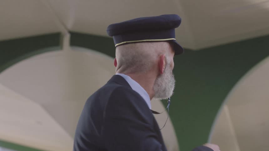 A distinguished, grey-bearded train conductor in uniform blows his whistle to signal departure at the station