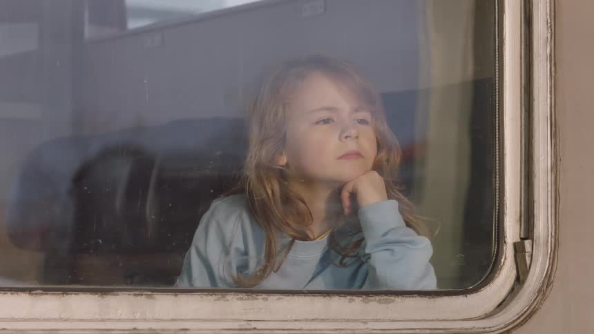 A thoughtful young girl with long hair looks out of a train window, resting her head on her hand