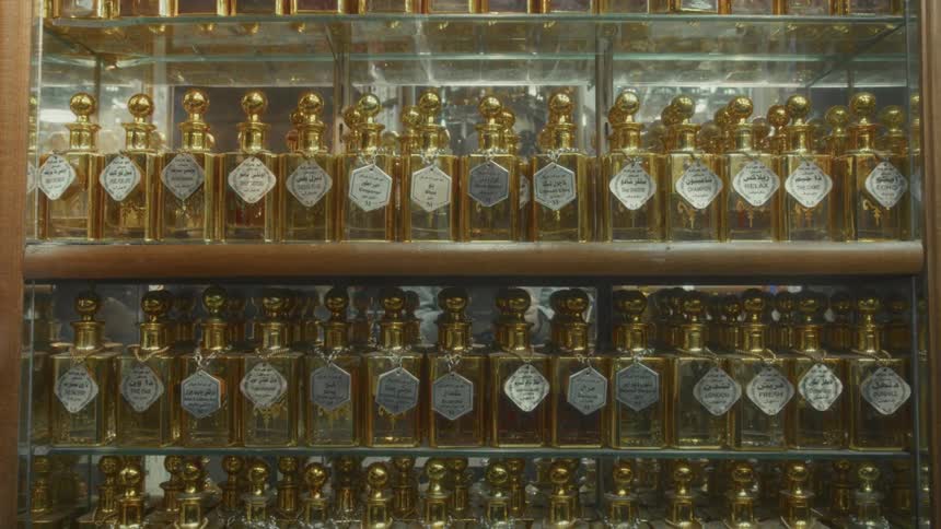 Rows of traditional Arabic perfume bottles with gold lids and silver labels are neatly displayed on shelves in a glass case