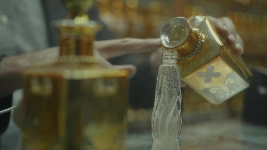 Close-up of a perfumer pouring a fragrance from a large ornate golden bottle into a smaller decorative glass vial