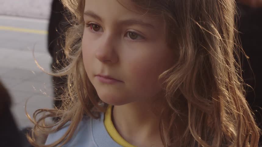 Close-up of a sad little girl's face, her golden hair blowing in the wind on a crowded train station platform