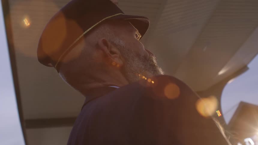 A thoughtful bearded captain in uniform stands silhouetted against a warm, flaring sun