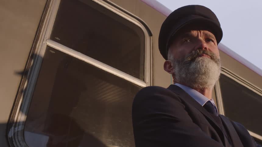 An elderly train conductor with a grey beard stands proudly next to a train car during golden hour