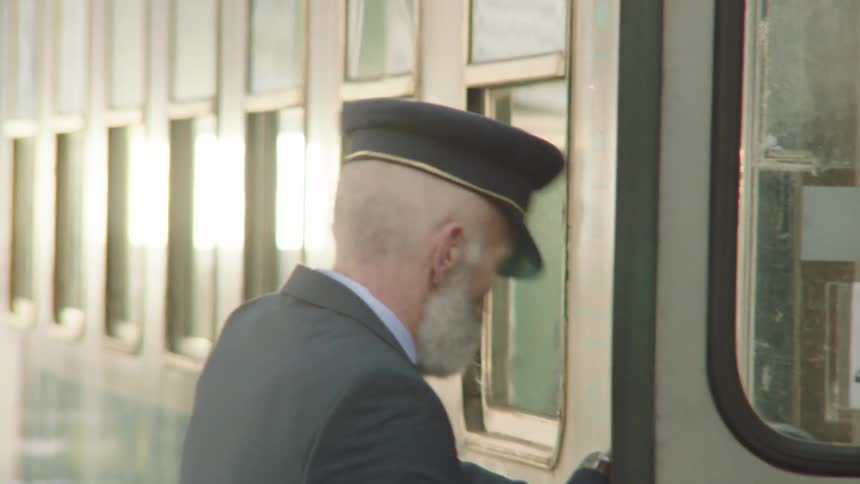 A senior train conductor with a white beard boards a train car at the station and closes the door