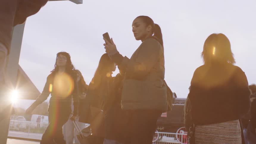 A mother and son look at a smartphone while waiting with a crowd of travelers at a sun-drenched train station
