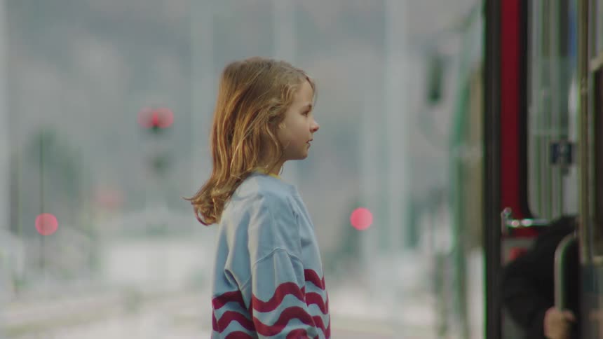 A young girl patiently waits on a platform as people board the train in slow motion