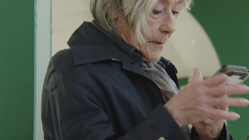 An elderly woman in a trench coat finishes a call and starts using her smartphone while waiting in public