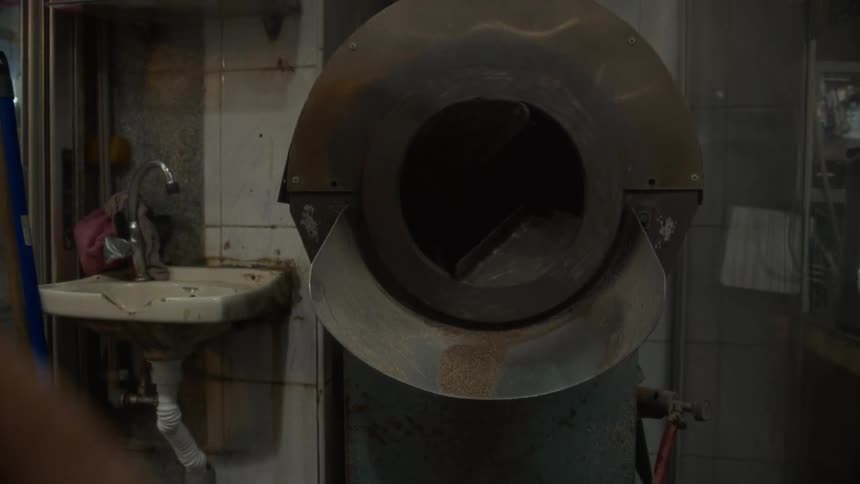 A worker scoops raw grains from a large metal tray and carefully pours them into an industrial roaster for processing