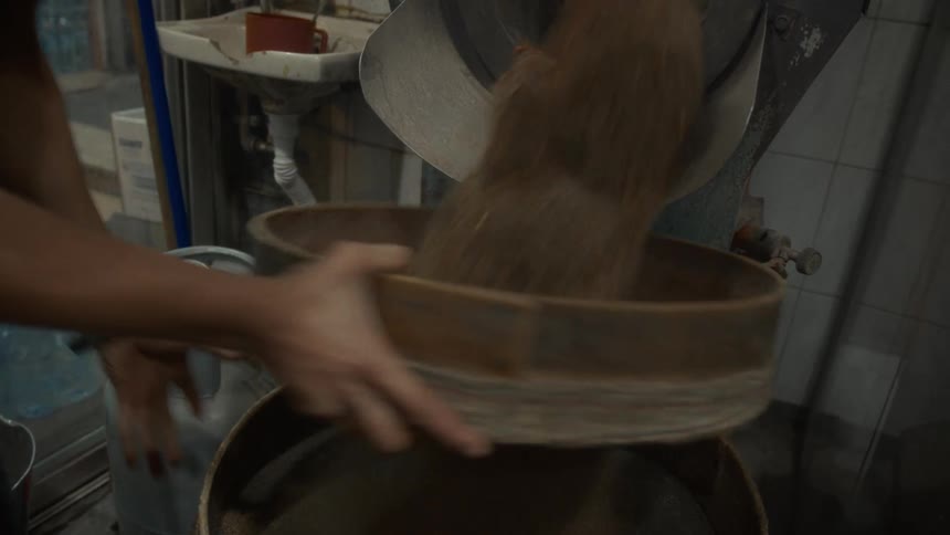 A worker manually sifts freshly roasted nuts and seeds through a large sieve to separate them from sand and dust