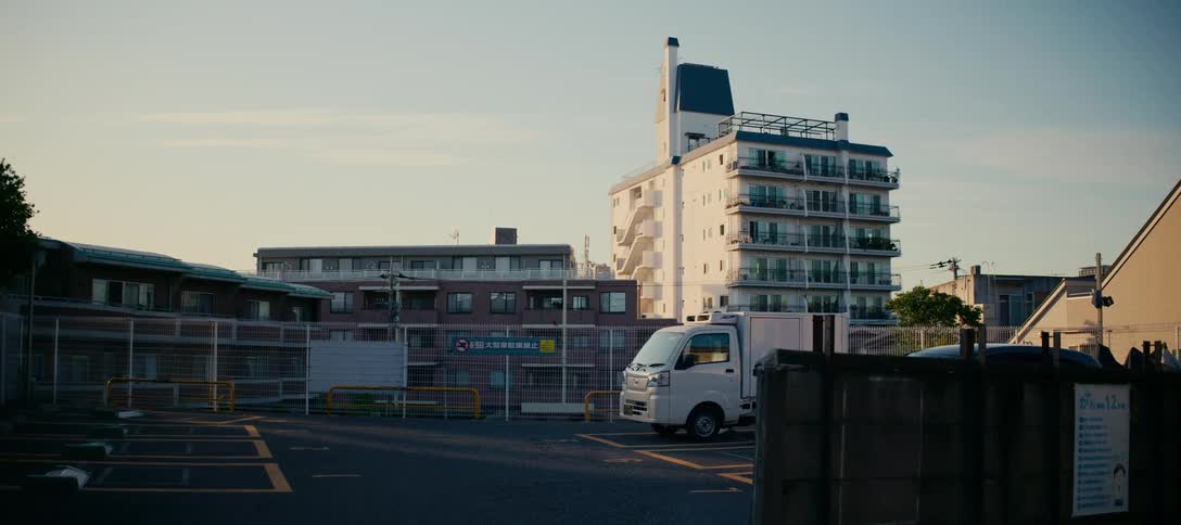 A quiet urban scene in Japan shows a small delivery truck parked in a lot surrounded by apartment buildings during sunset