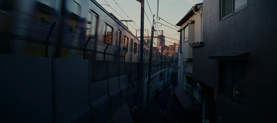 A passenger train travels on an elevated track through a narrow, shadowy backstreet in a Tokyo neighborhood at sunset