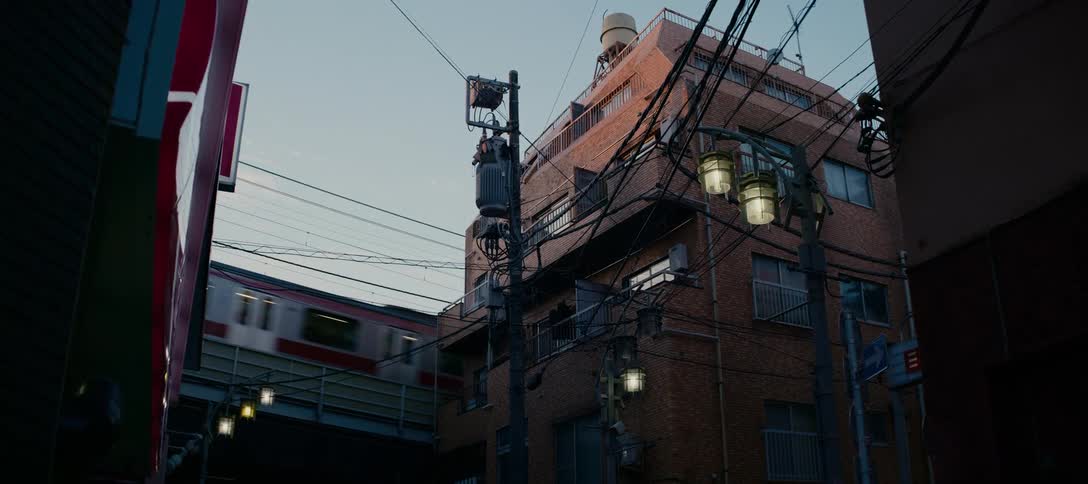 An elevated commuter train passes by a brick building and tangled power lines in a quiet urban alley in Tokyo, Japan at sunset