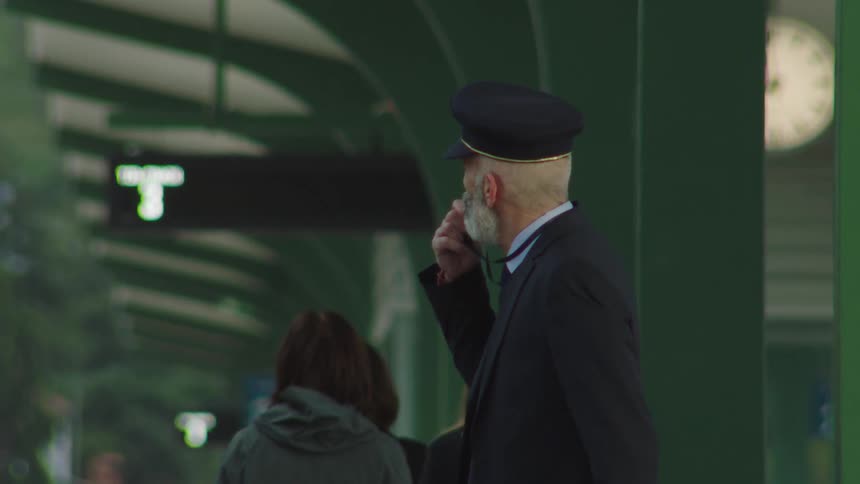 A senior train conductor with a distinct white beard stands on a platform and blows his whistle to signal departure