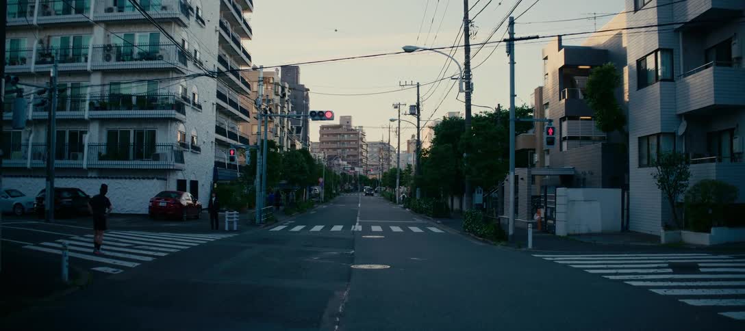 A quiet residential street in a Japanese city is empty except for a few people waiting at a crosswalk during the golden hour