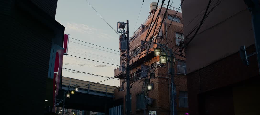 A low-angle shot captures a brick apartment building and a complex web of power lines on a quiet urban street in Japan during sunset