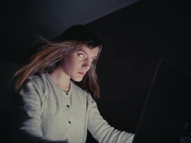 A woman with unsettling white eyes stares intently at her laptop screen in a dark room