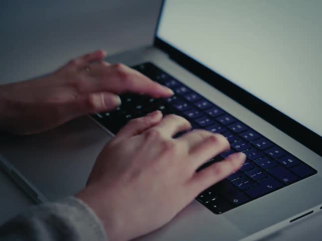 Close-up of hands typing on a laptop keyboard in the dark, illuminated by the screen's glow