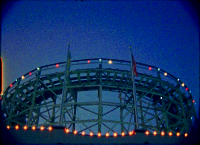 A grainy, vintage 8mm film shot captures the 'Dragon Coaster' ride lit up with colorful lights against the night sky