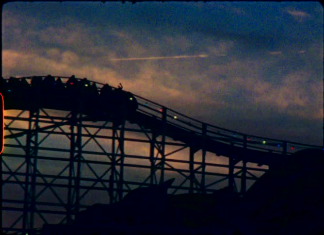 Vintage film footage captures the silhouette of a wooden roller coaster climbing slowly against a cloudy twilight sky