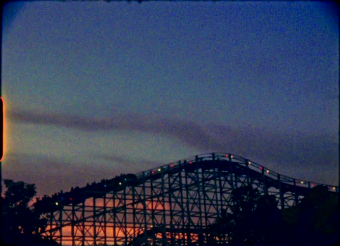 Vintage 8mm footage shows a wooden roller coaster with colorful lights climbing a hill against a beautiful sunset sky