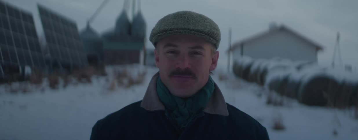 A proud farmer with a mustache and flat cap offers a gentle smile while standing on his snow-covered farm