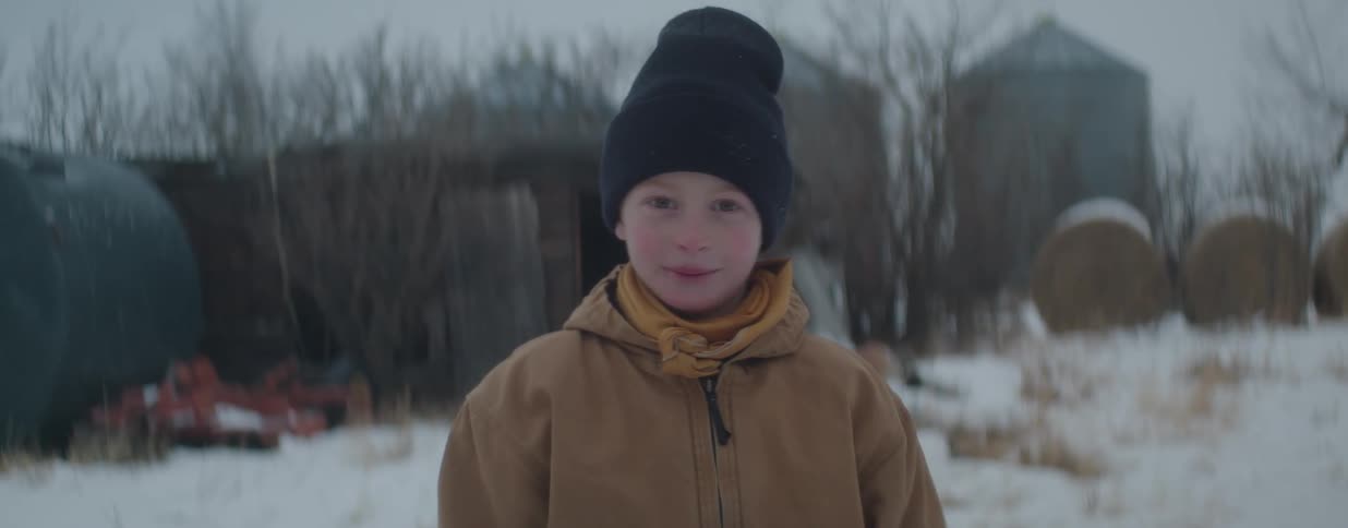 A close-up portrait of a young boy wearing a warm winter coat and beanie while standing in the snow on a rural farm